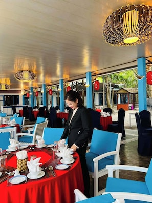 A restaurant with round tables covered in red tablecloths, blue chairs, and a waiter attending to a table. - Lang Co Beach Resort (Phu Loc)