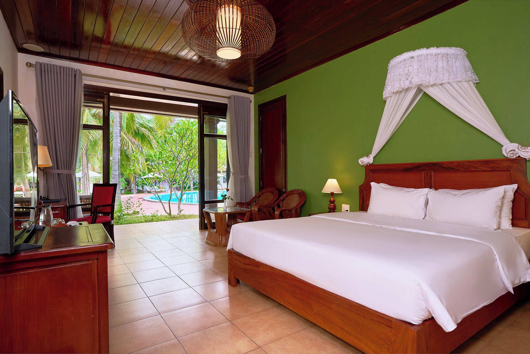 A hotel room with a large bed, wooden furniture, a desk, and a view of a pool and greenery.
