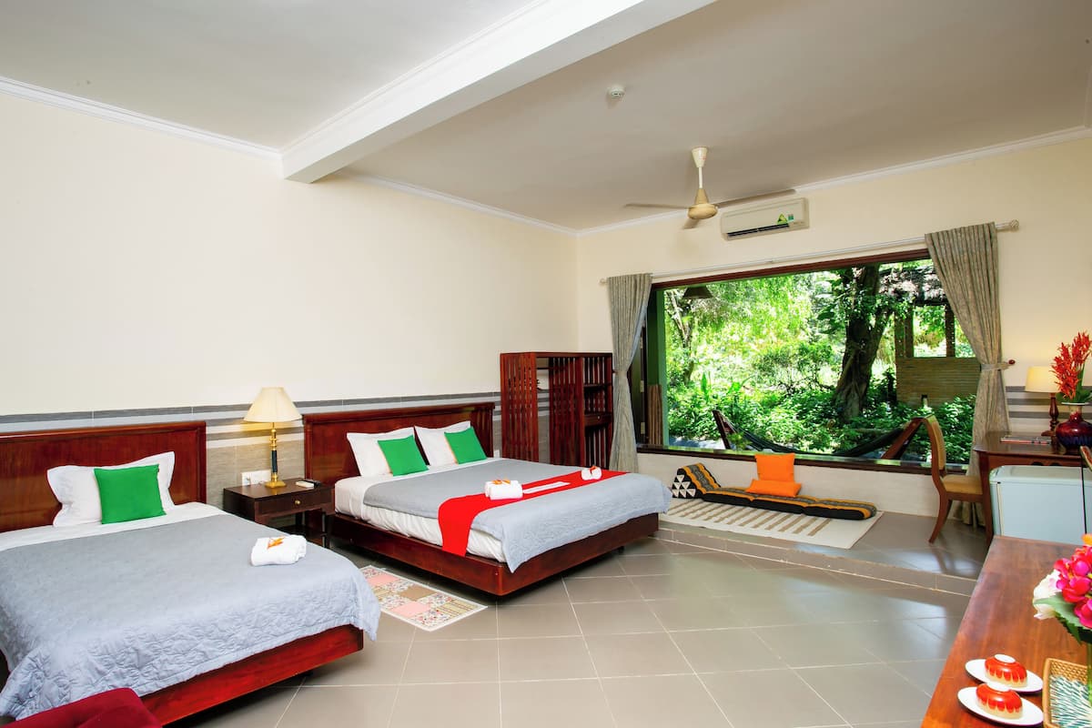 A hotel room with two beds, a desk, a chair, a window with a view of greenery, and a ceiling fan.
