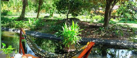 A hammock with a table and a potted plant, set by a pond in a lush garden.