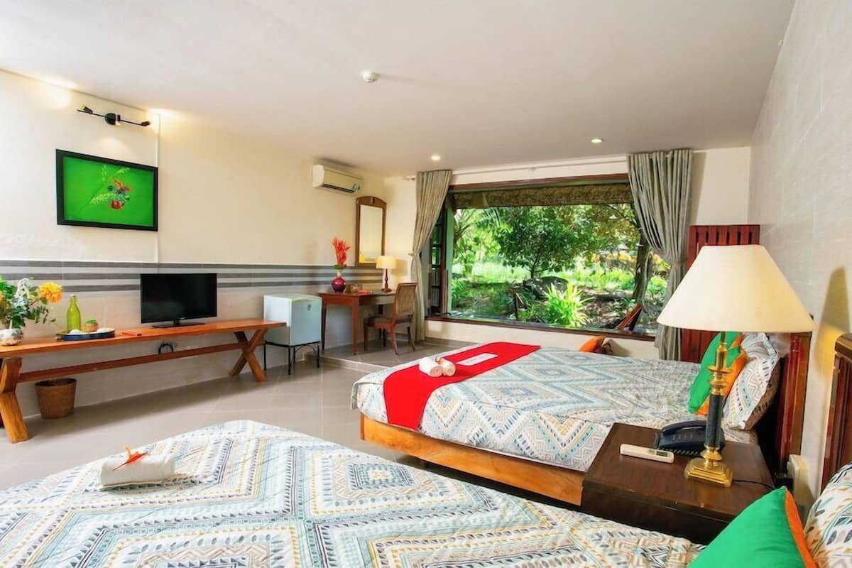 A hotel room with two beds, a desk, a TV, and a large window with a view of greenery.