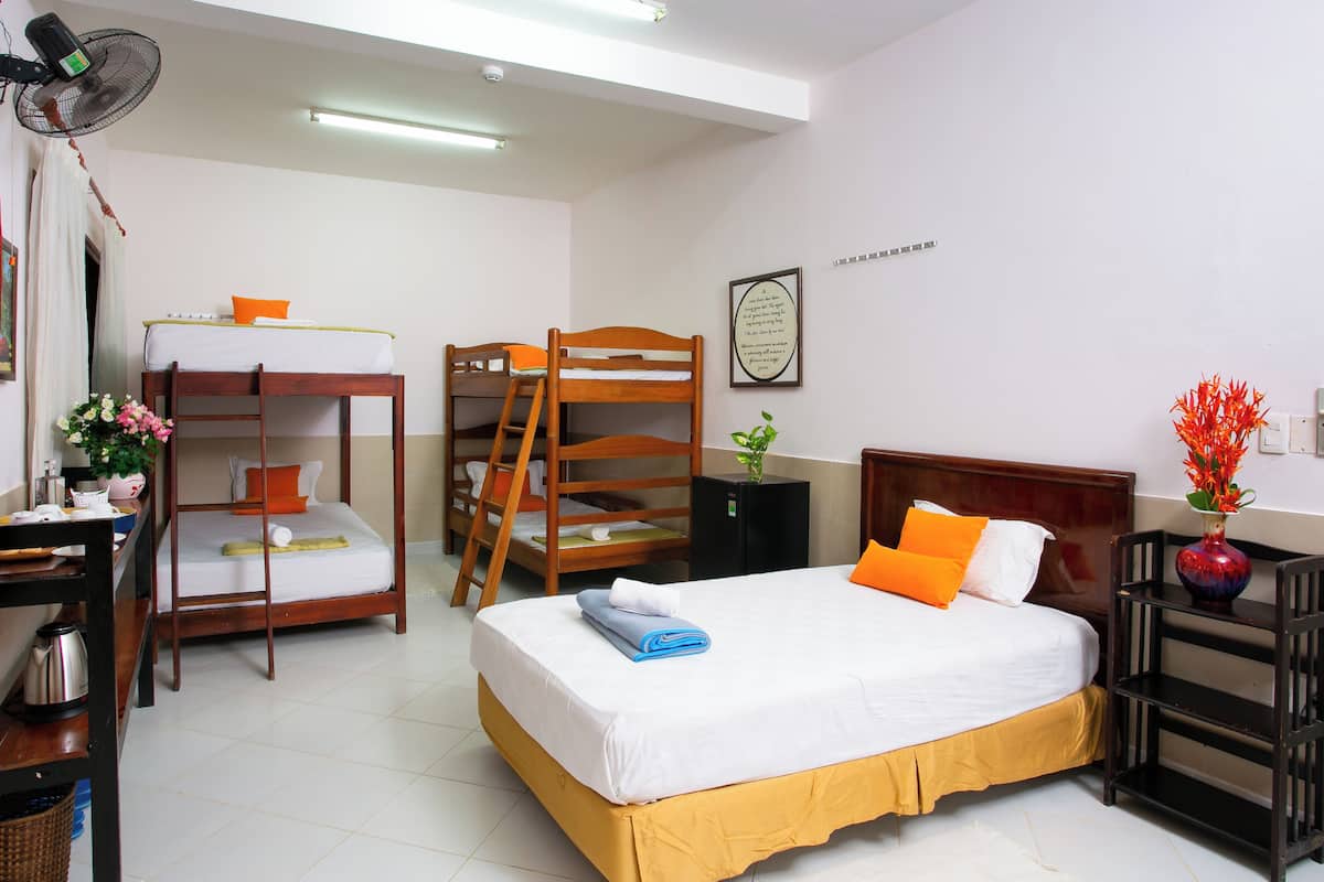 A hotel room with bunk beds, a desk with a fan, a small table with a kettle, and a bedside table with a vase of flowers.