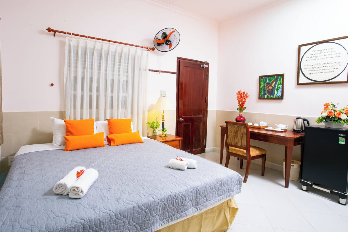 A hotel room with a bed, a desk, a chair, a small refrigerator, a fan, a door, and a framed quote on the wall.