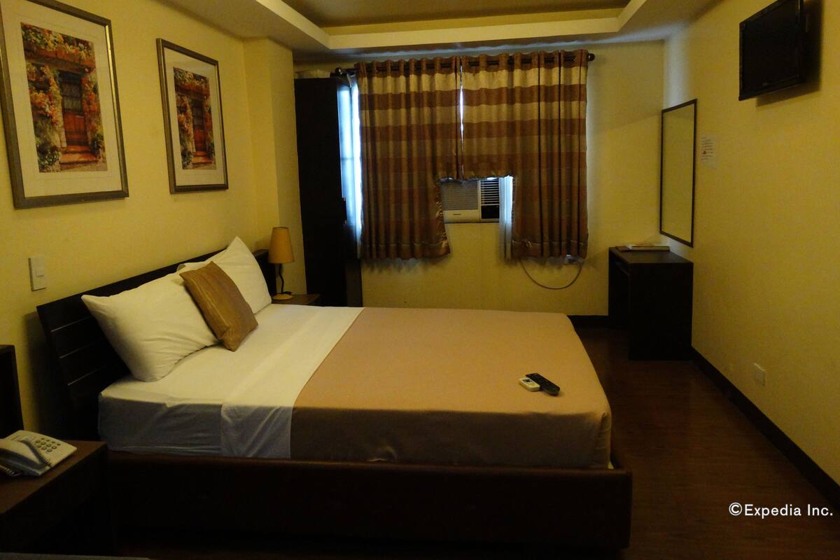 Habitación de hotel con una cama grande, una mesita de noche con lámpara, un televisor en la pared y un equipo de aire acondicionado.
