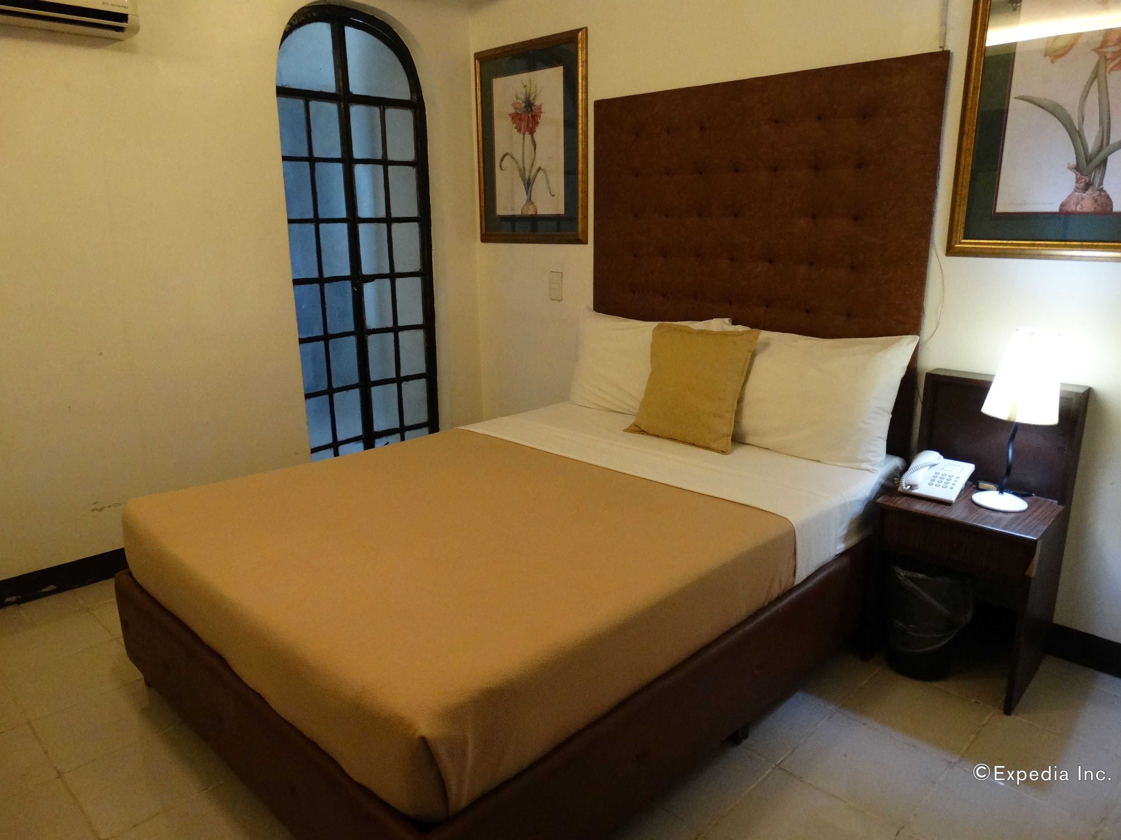 Camera d'albergo con un letto grande, un comodino con una lampada, un cestino e due quadri appesi al muro.