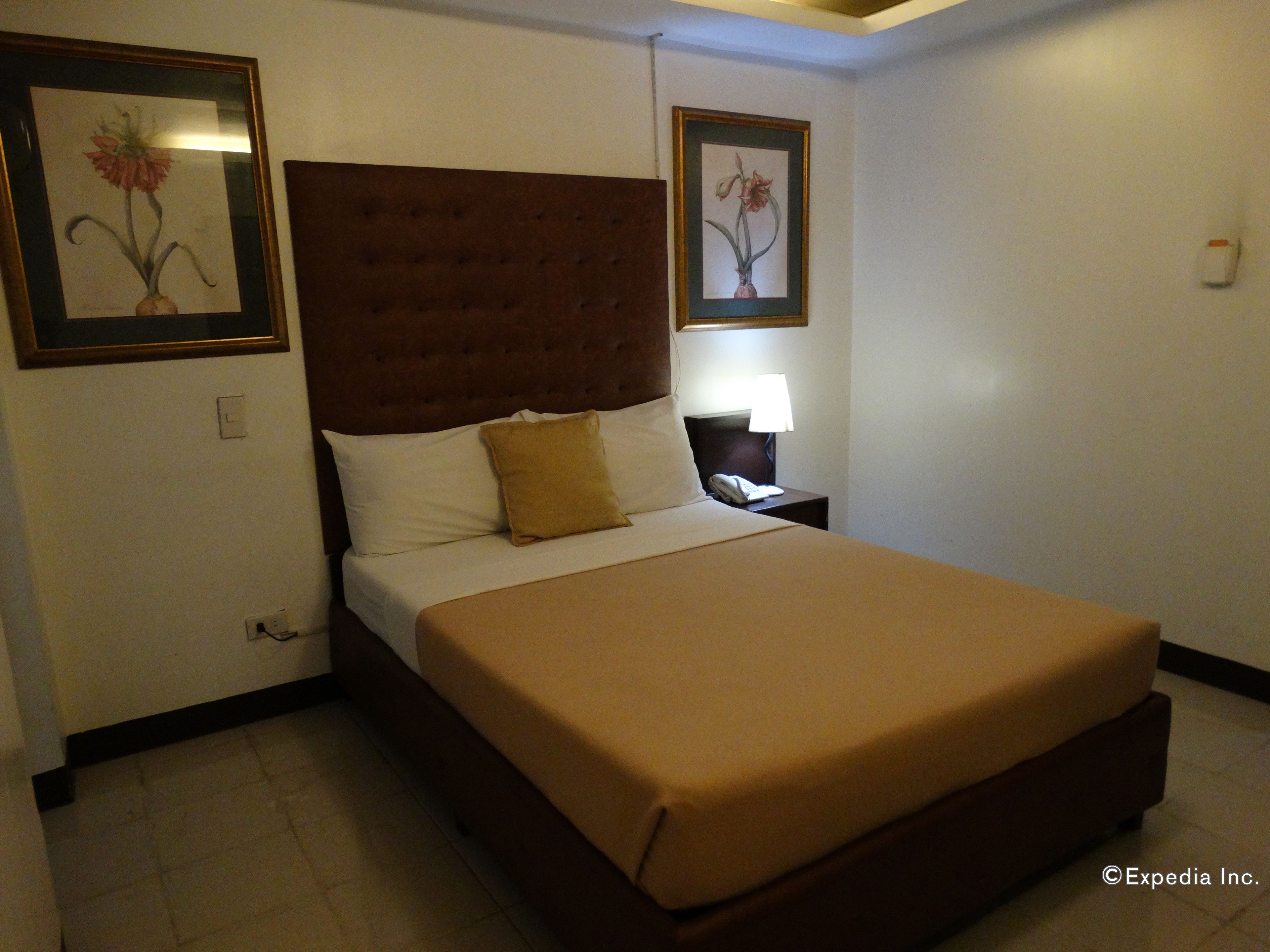 Un cuarto de hotel con una cama grande, una mesita de noche con lámpara, y dos cuadros enmarcados con motivos florales en las paredes.