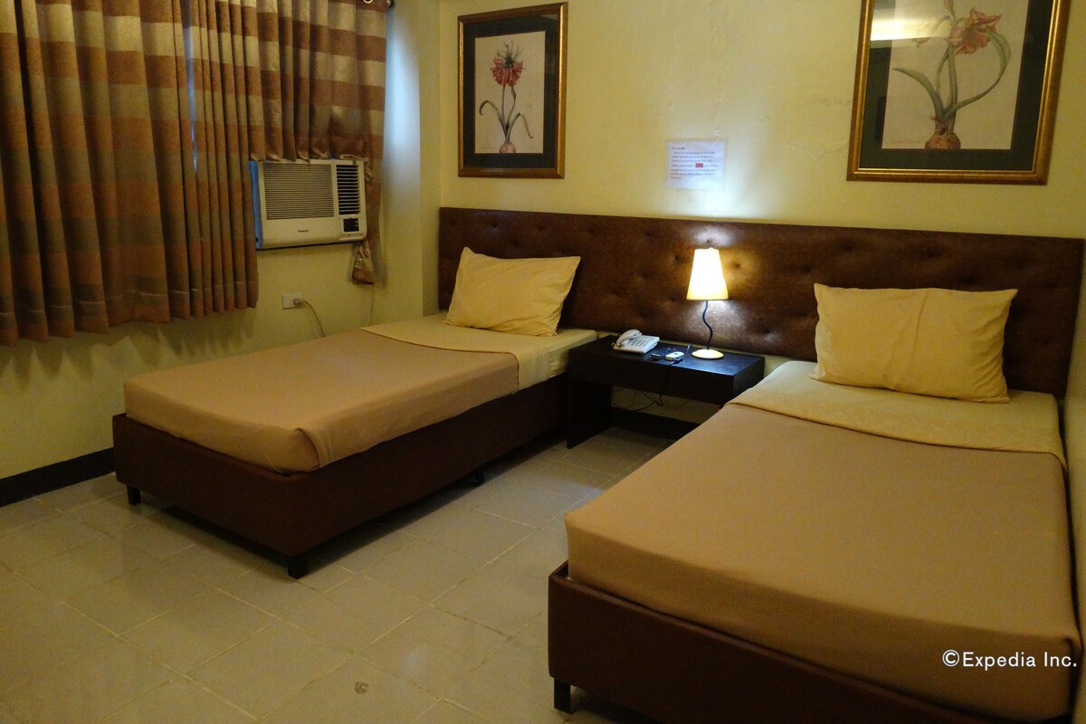 A hotel room with two single beds, a nightstand with a lamp, a telephone, and a framed picture on the wall.
