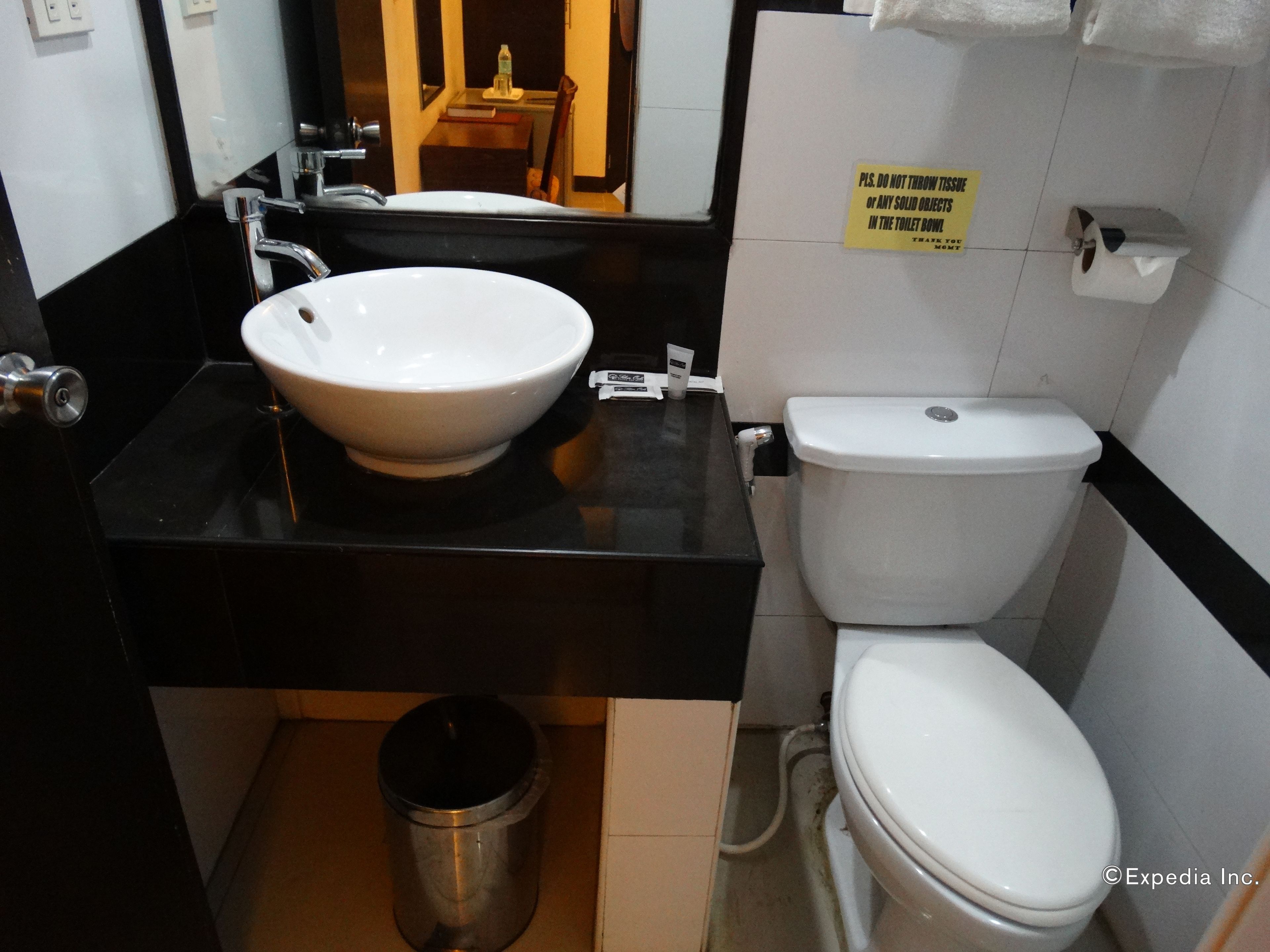 Une salle de bain avec un lavabo blanc, un plan de toilette noir et des toilettes. On trouve une poubelle sous le lavabo et un panneau sur le mur.
