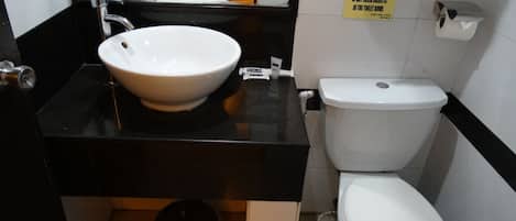 Un baño con lavamanos blanco, encimera negra y un inodoro. Hay un cubo de basura debajo del lavamanos y un cartel en la pared.