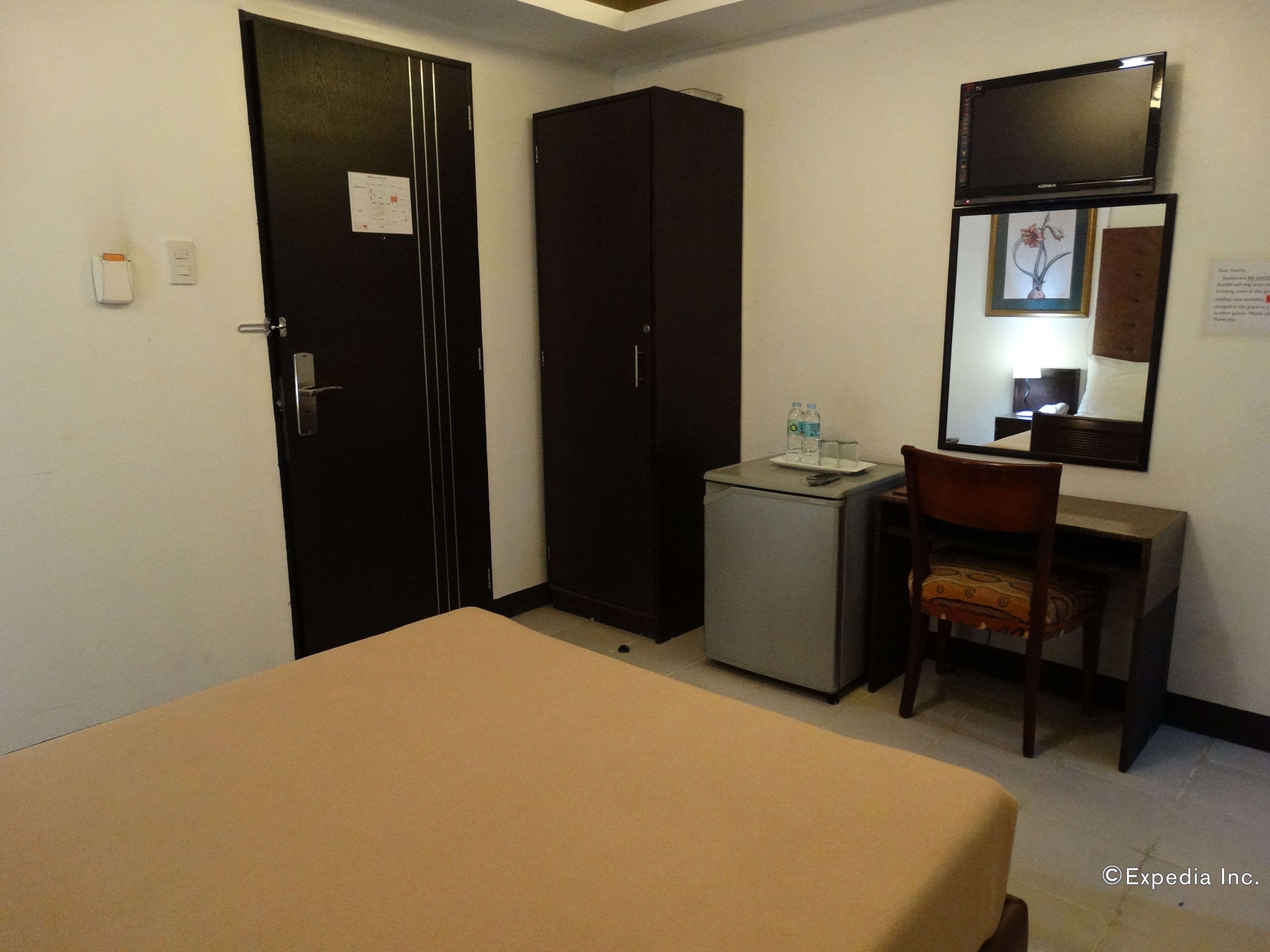 A hotel room with a bed, a desk, a chair, a small refrigerator, and a mirror on the wall.