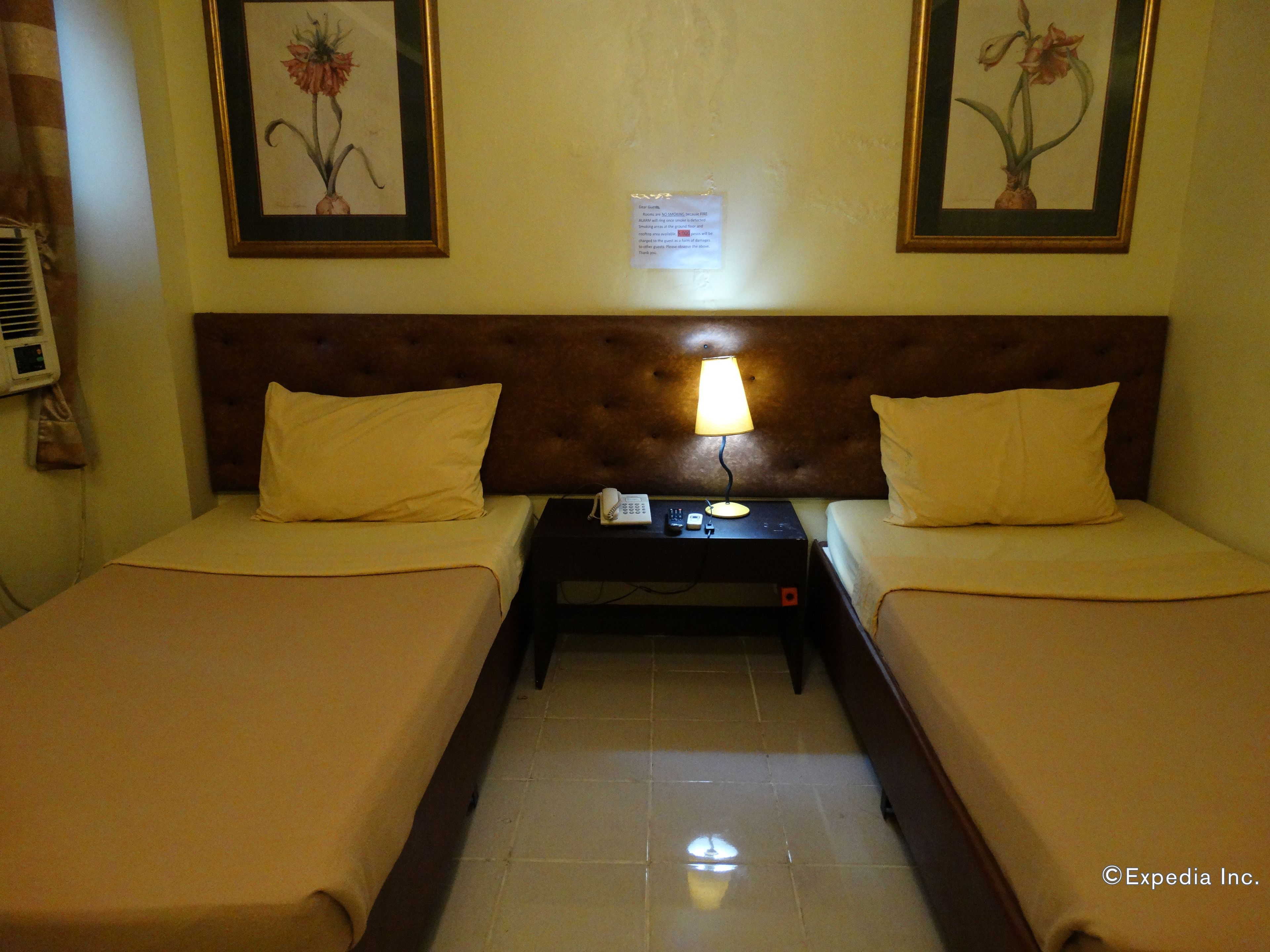 A hotel room with two single beds, a nightstand with a lamp, a telephone, and framed floral artwork on the walls.