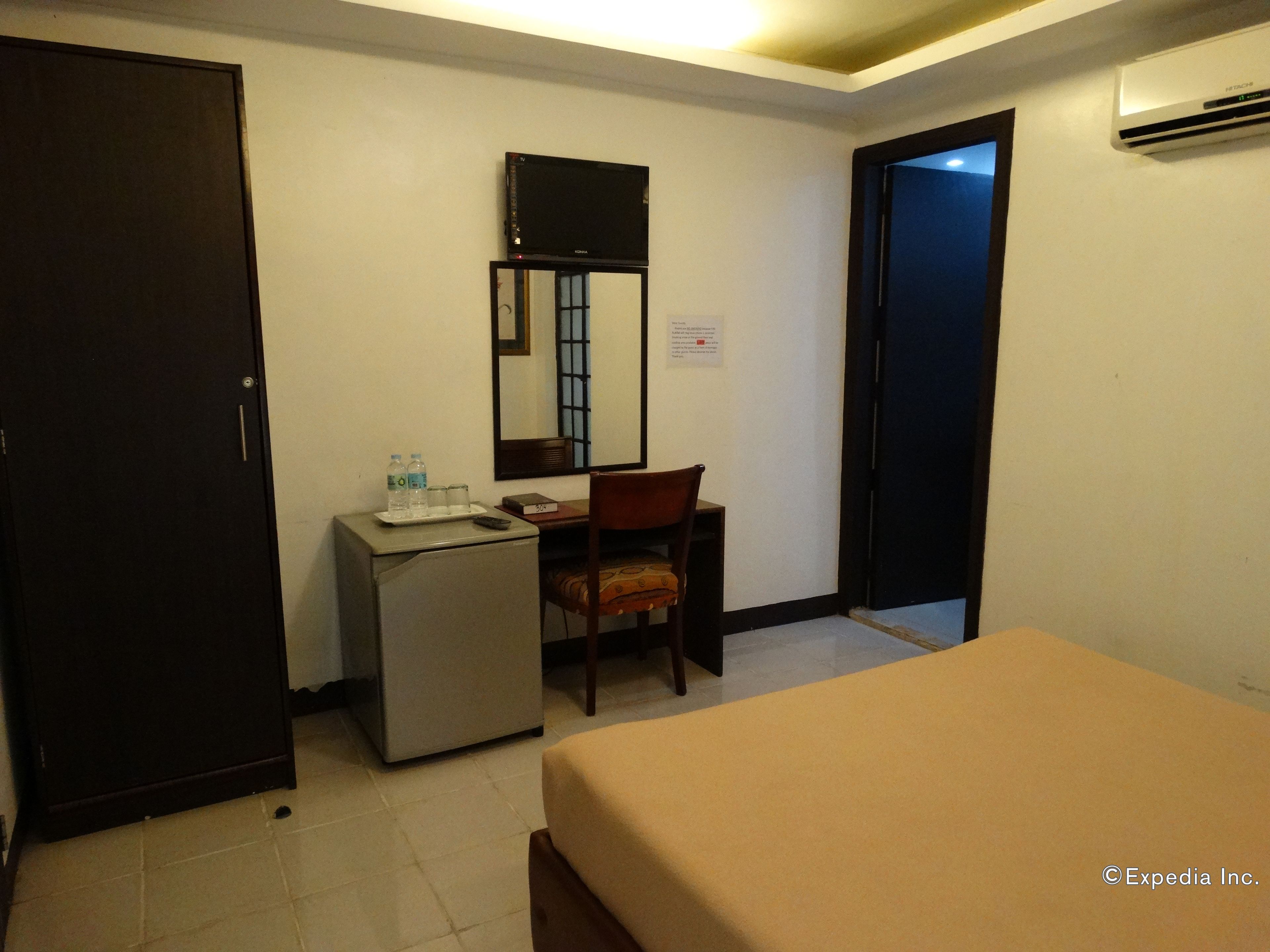 A hotel room with a bed, a desk with a chair, a small refrigerator, and a mirror on the wall.