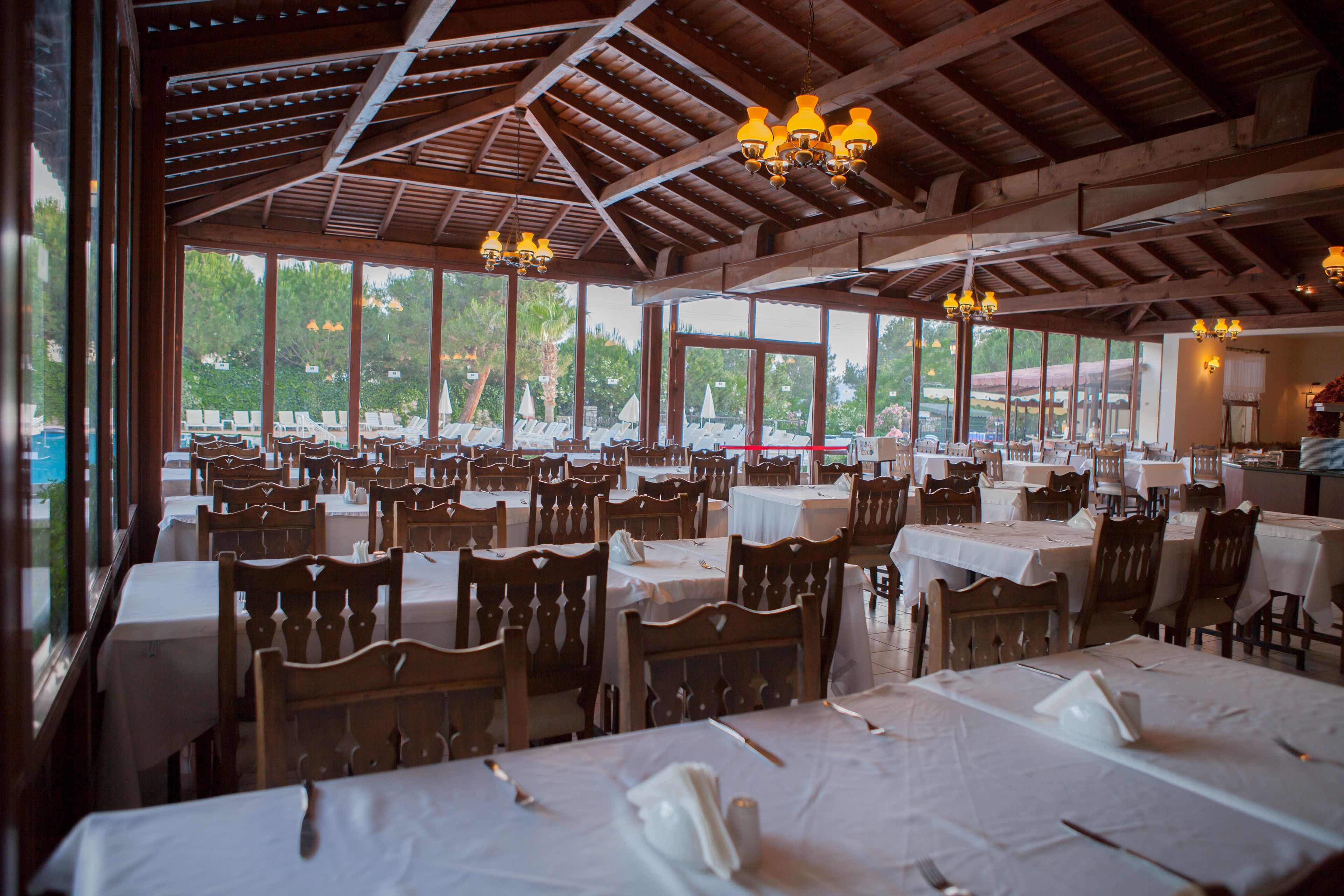 Un vaste espace dédié à la salle à manger, avec de nombreuses tables et chaises, de grandes fenêtres offrant une vue sur l’extérieur et un plafond en bois agrémenté de luminaires suspendus.