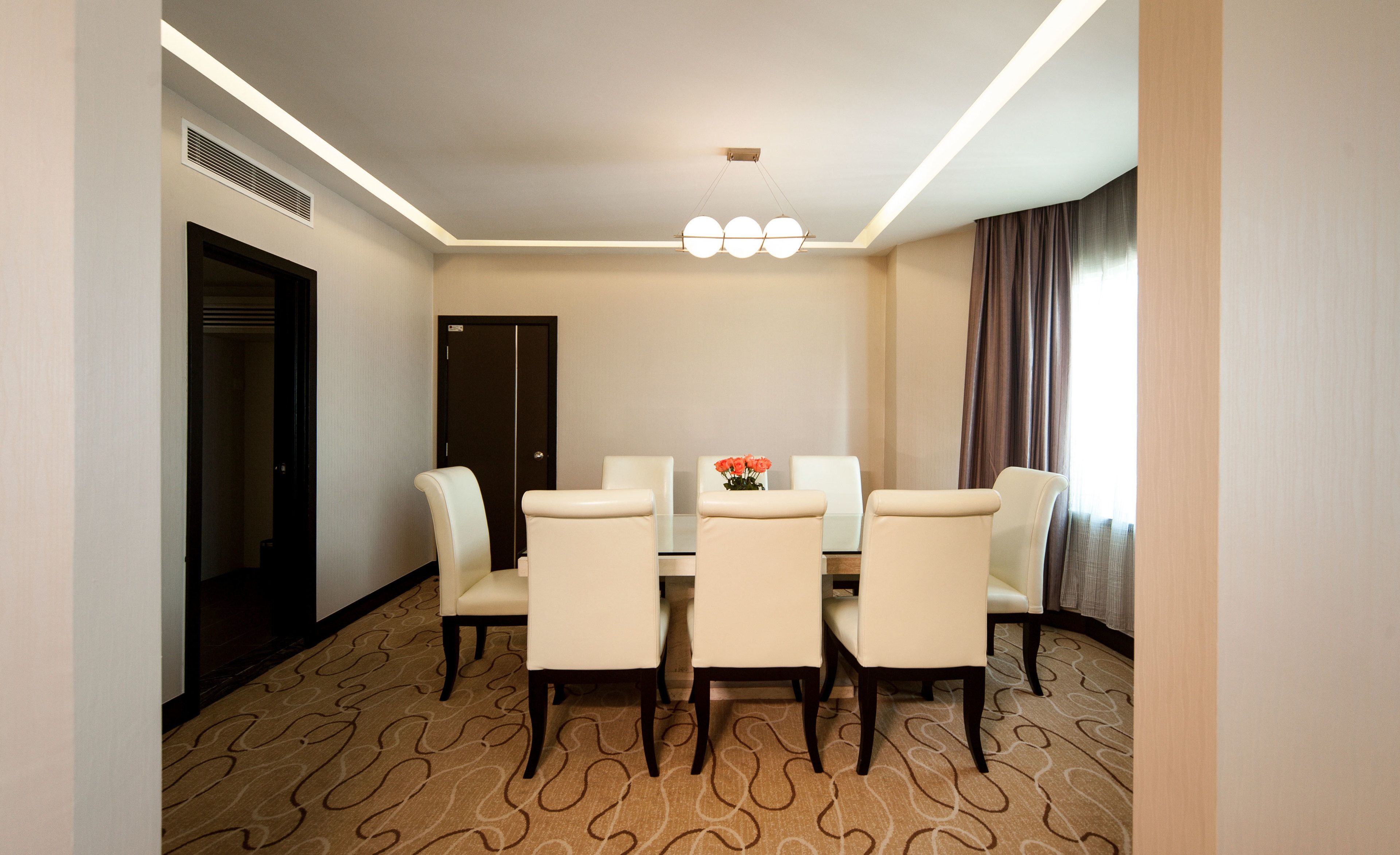 A hotel dining room with a rectangular table, six chairs, a vase with flowers, and a large window with curtains.
