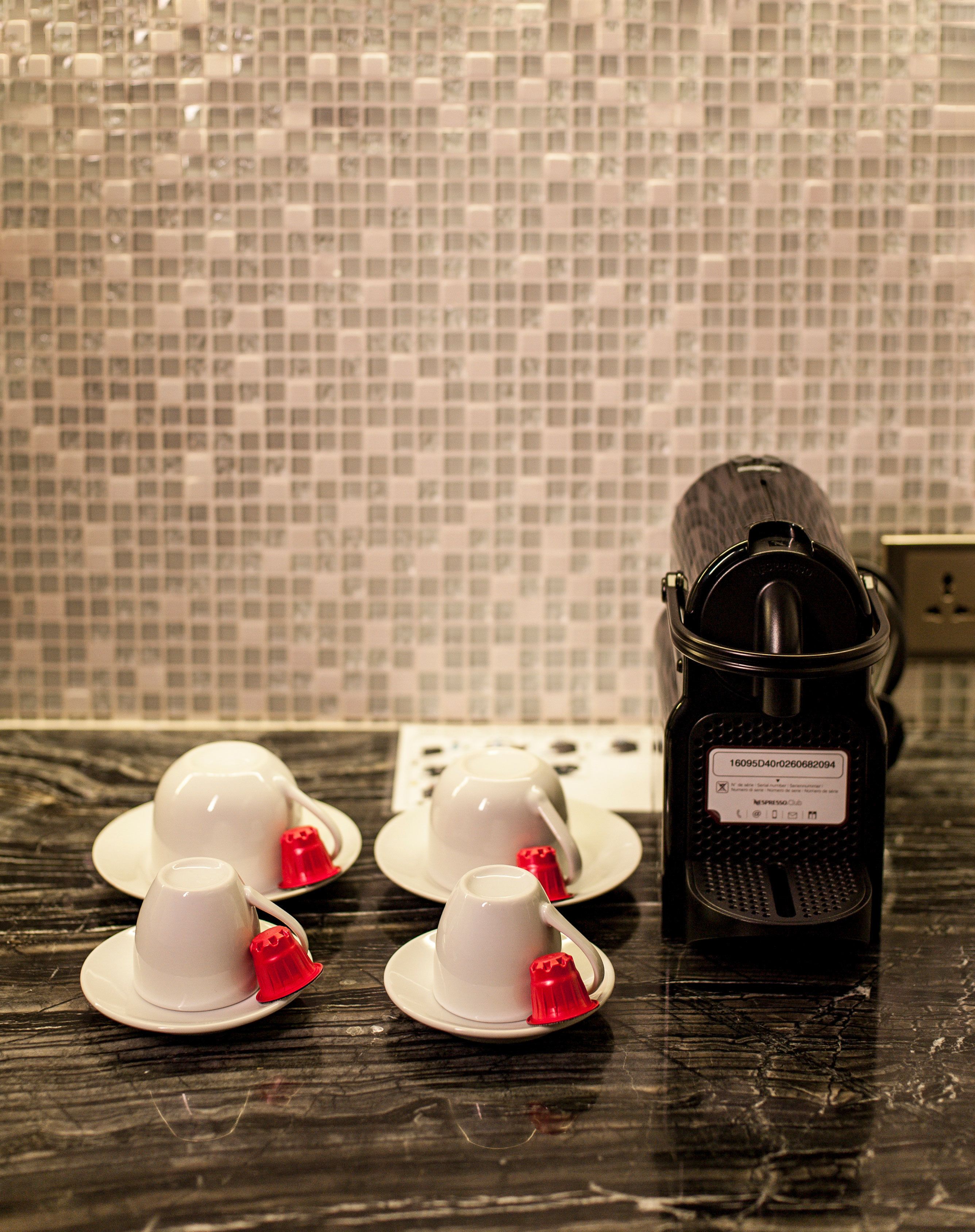 A kitchen area with a coffee machine, a tiled backsplash, and three white cups with red lids on a dark countertop.
