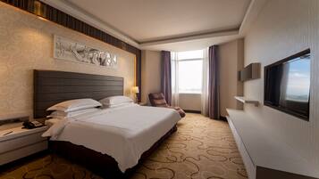 A hotel room with a large bed, a flat-screen TV, a chair, and a desk with a phone.