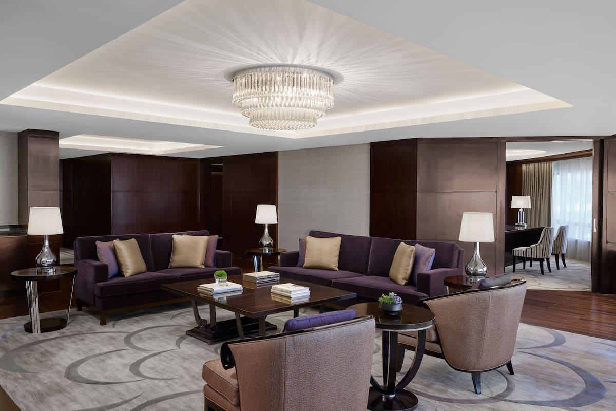 A modern hotel lobby with a large chandelier, dark wood paneling, a seating area with sofas and armchairs, and a dining area in the background.