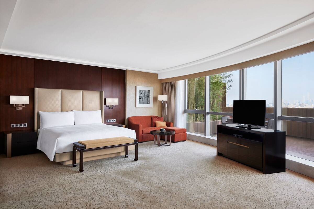 A hotel room with a large bed, a sofa, a TV, and a balcony with a city view.