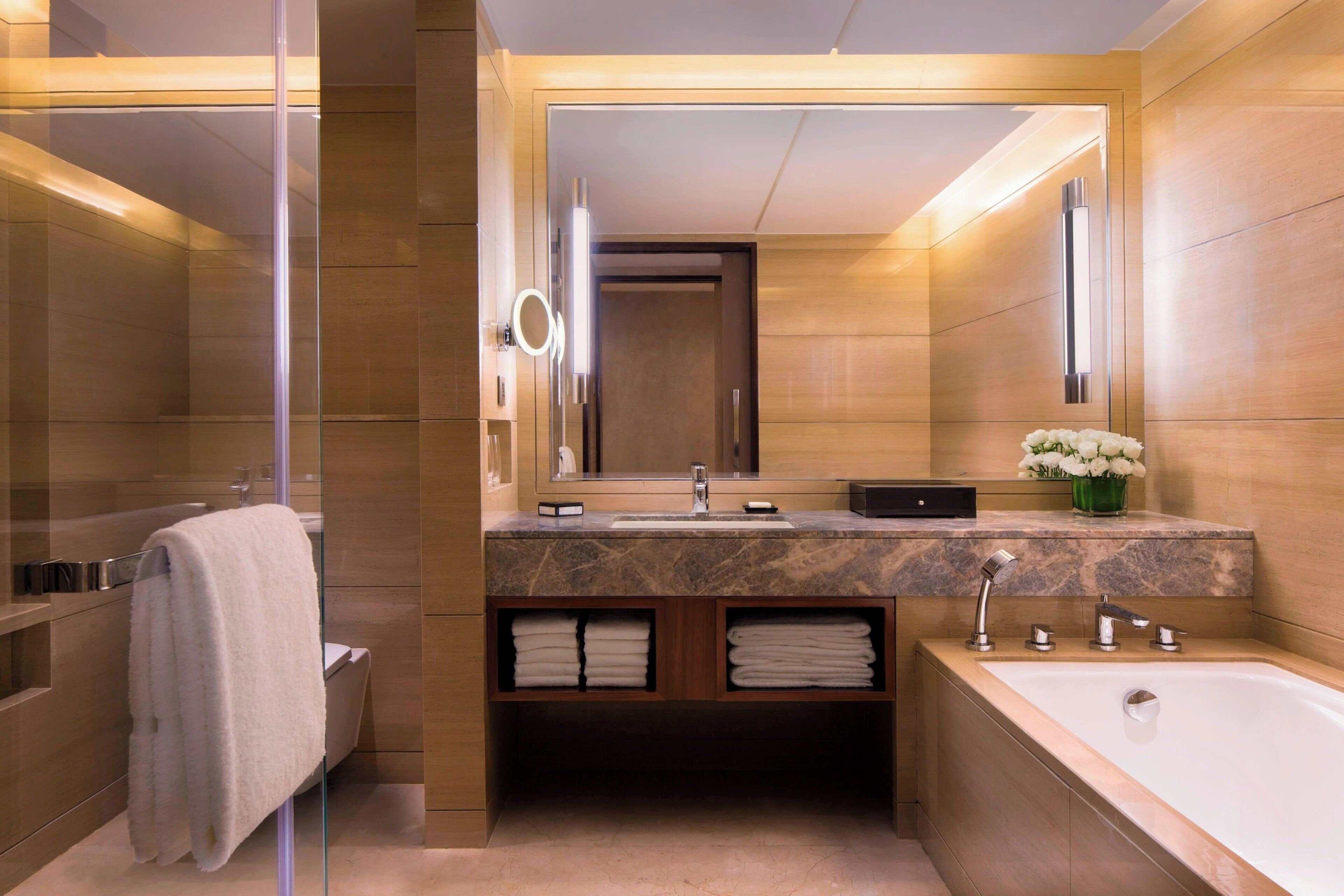 A modern bathroom with a glass shower enclosure, a marble vanity with a large mirror, a bathtub, and a green vase with flowers.