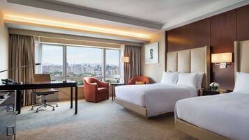 Habitación de hotel con escritorio, dos camas, una silla, televisor y vistas a la ciudad.