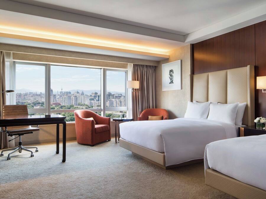 Habitación de hotel con escritorio, dos camas, una silla, televisor y vistas a la ciudad.