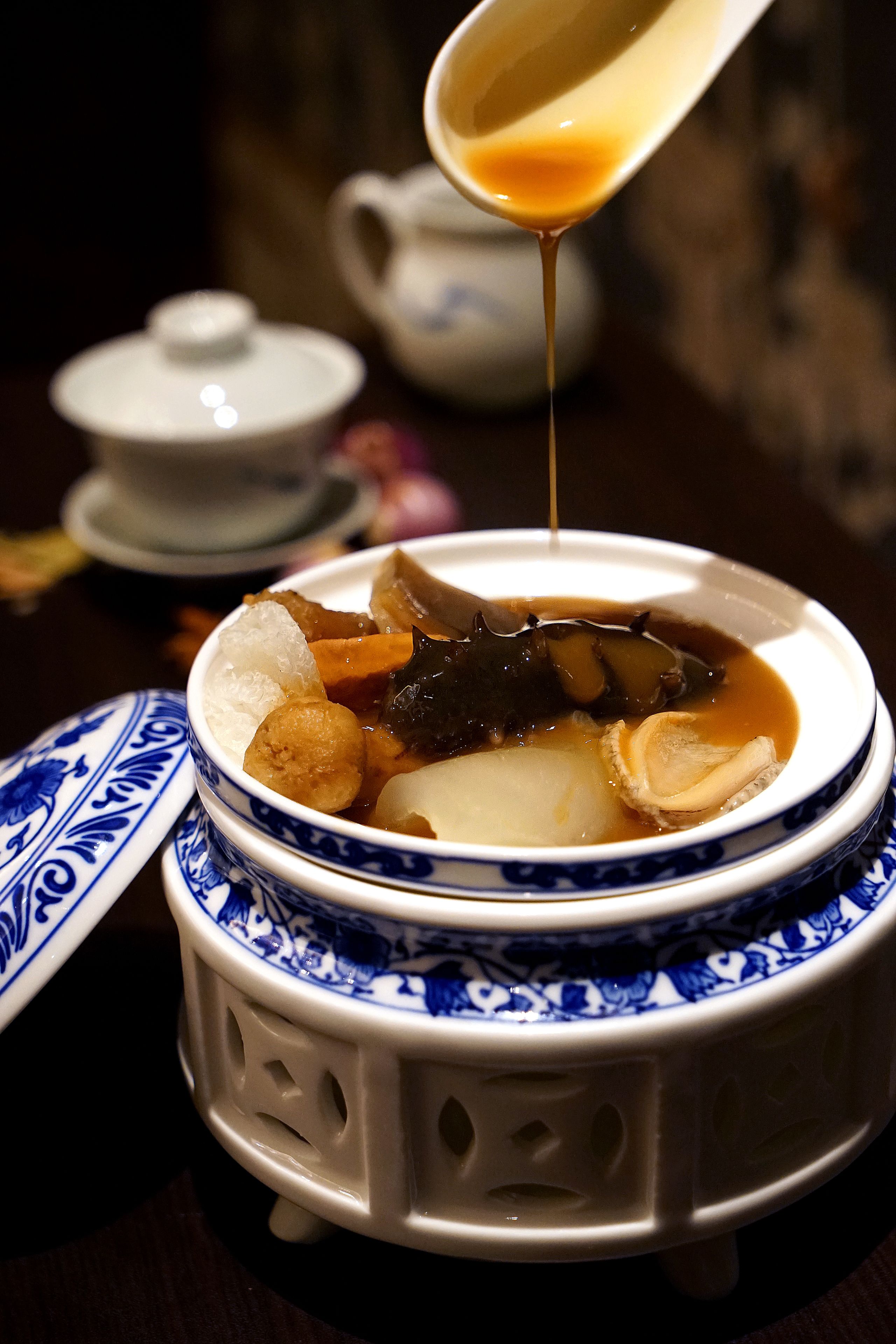 A traditional ceramic bowl with a lid, containing a soup with various ingredients, and a spoon pouring a liquid into it.