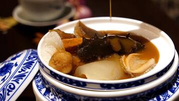 A traditional ceramic bowl with a lid, containing a soup with various ingredients, and a spoon pouring a liquid into it.