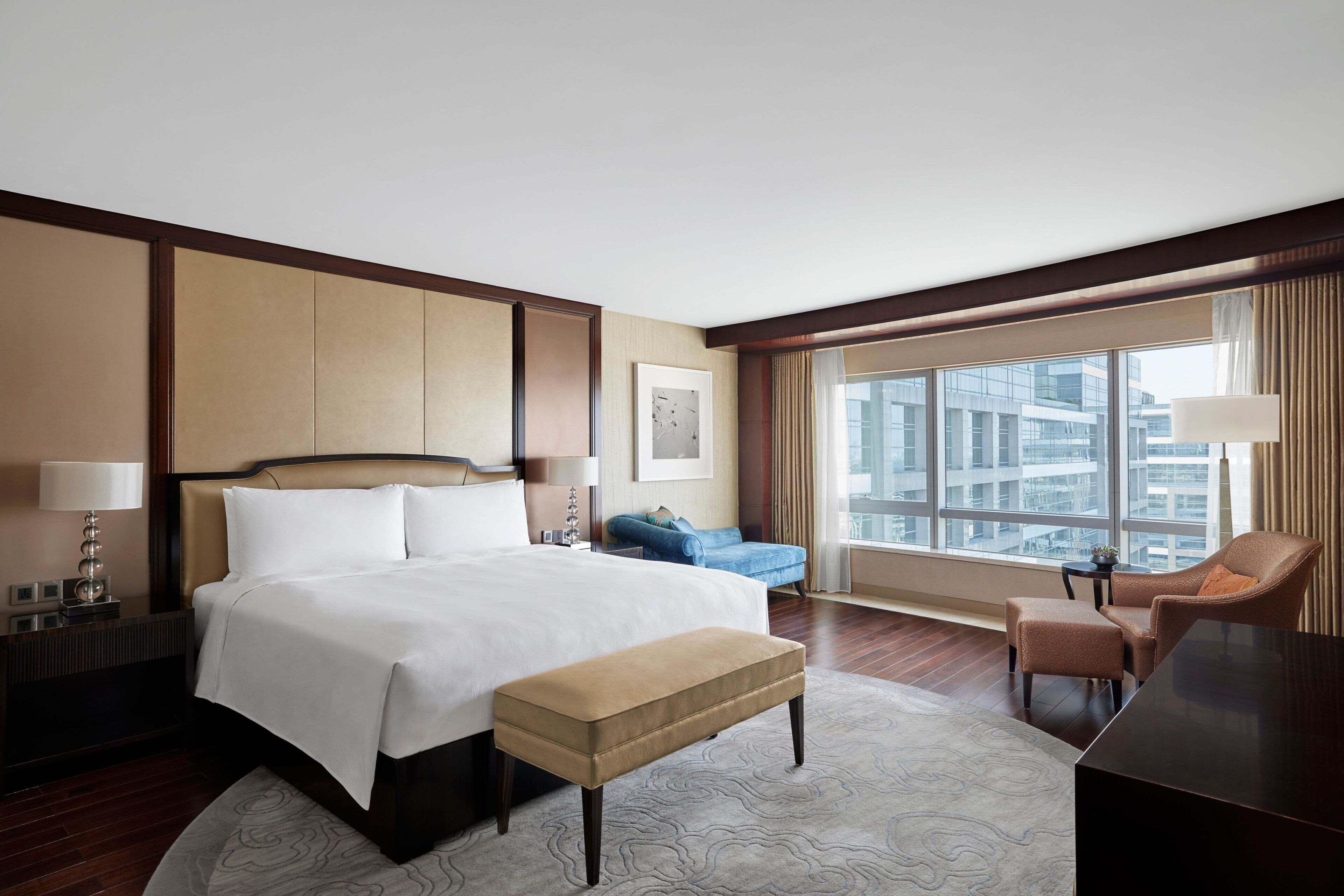 A modern hotel room with a large bed, a desk, a chair, and a view of the cityscape through large windows.