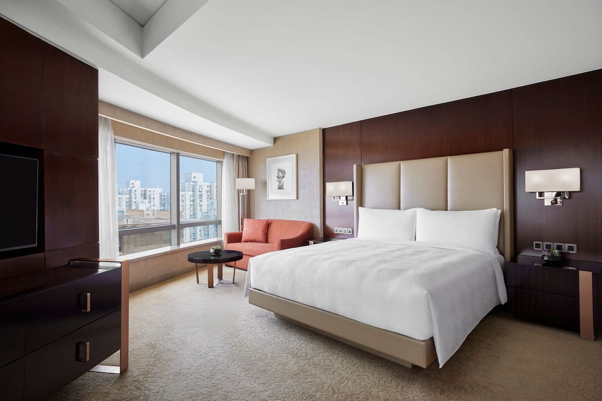 A modern hotel room with a large bed, a desk, a chair, a TV, and a view of the city.