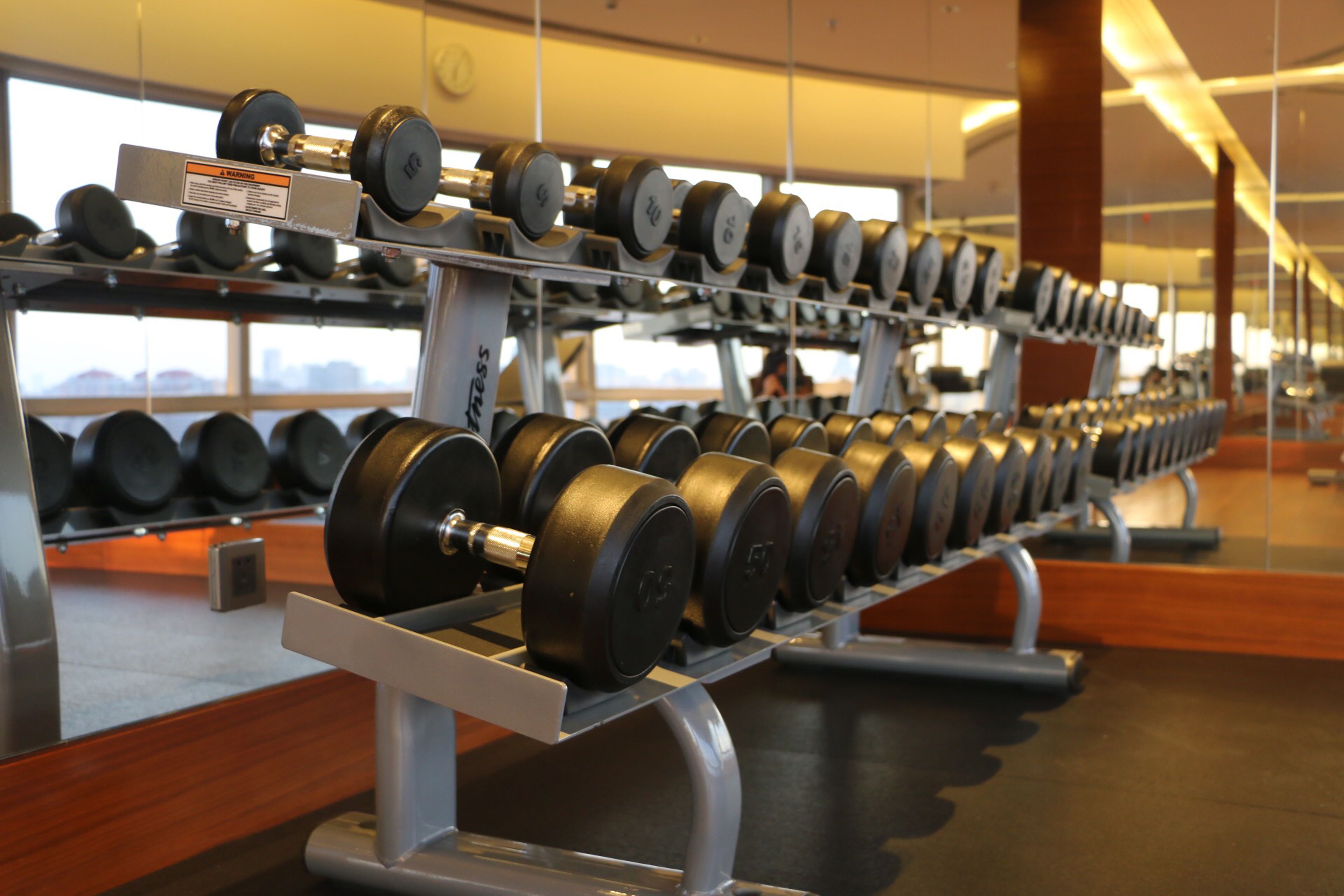 A gym with rows of dumbbells on racks, a wooden floor, and large windows in the background.