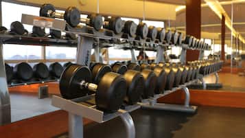 A gym with rows of dumbbells on racks, a wooden floor, and large windows in the background.