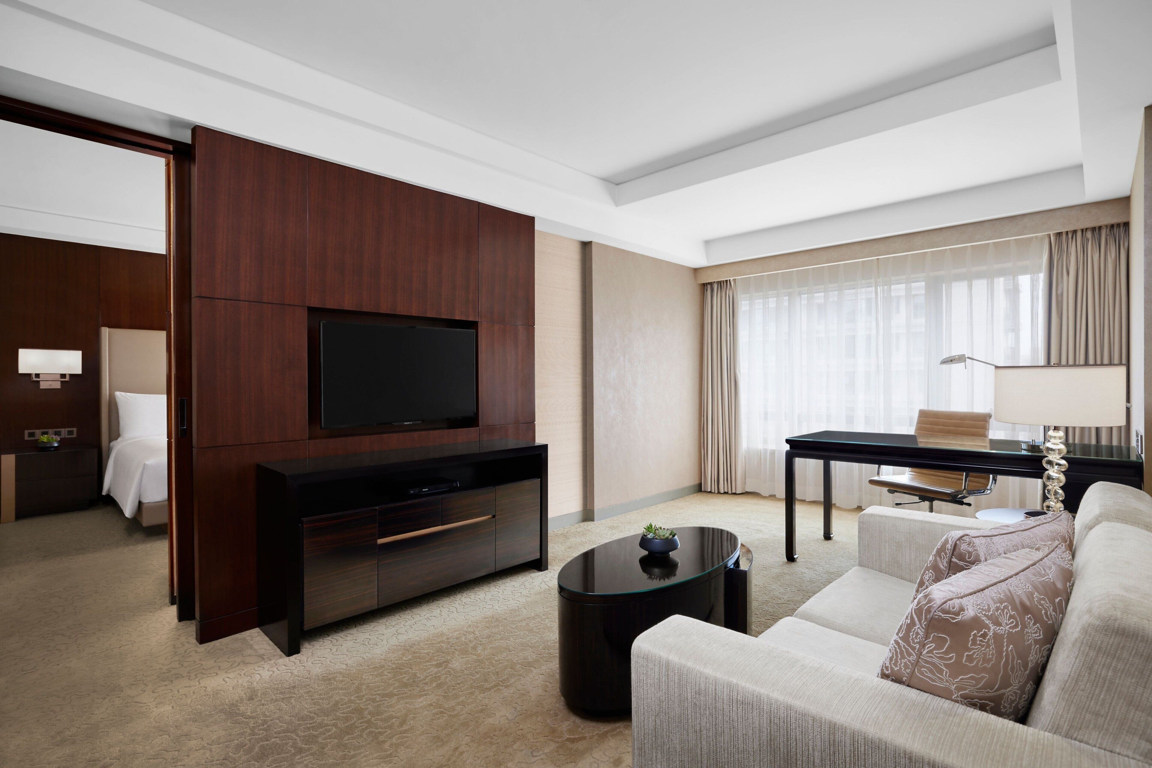 A modern hotel room with a flat-screen TV, a sofa, a coffee table, and a desk with a lamp.