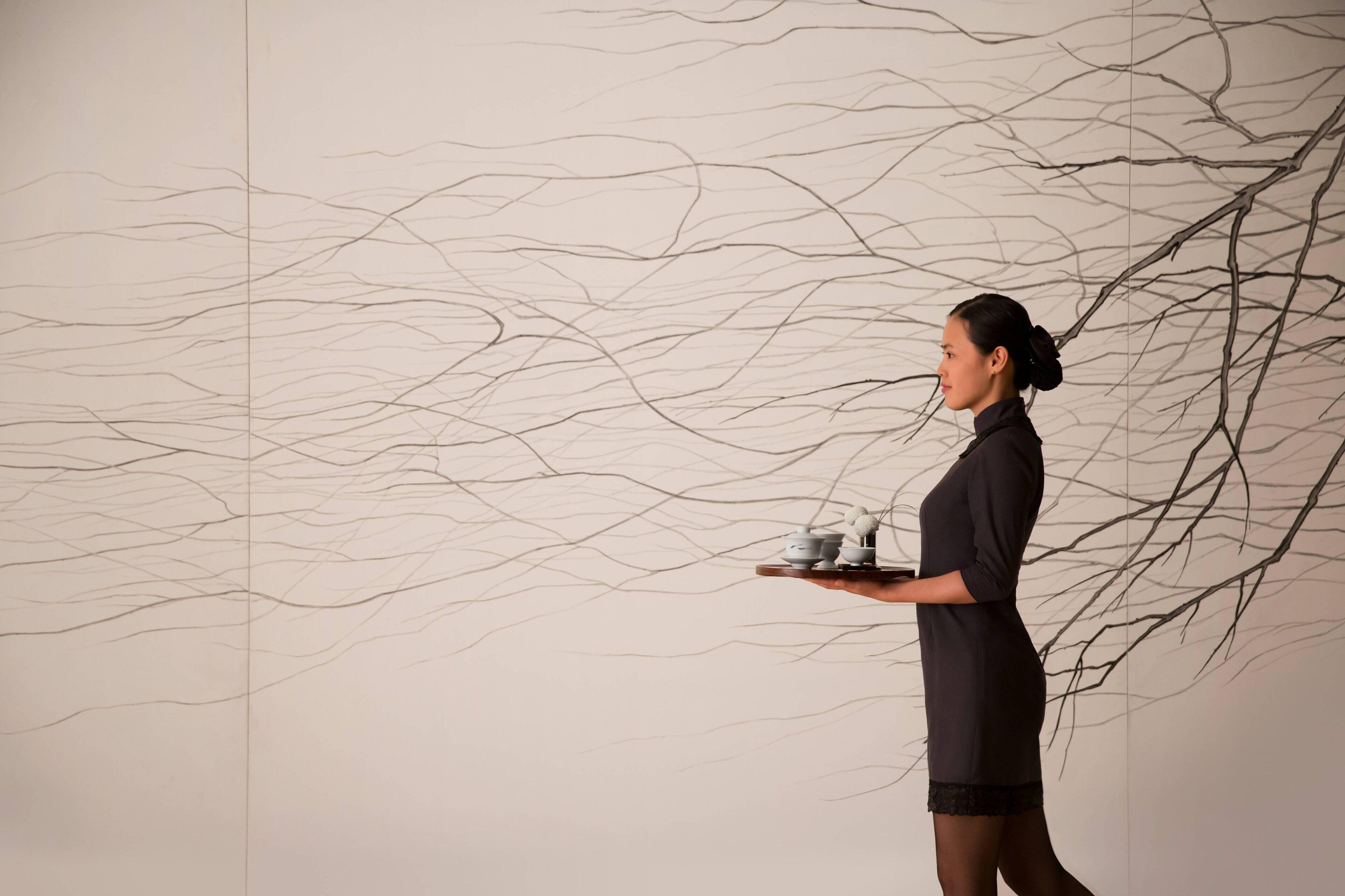 A person holding a tray with a teapot and cups, standing in front of a wall with abstract line art.
