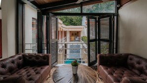 Une pièce dotée de deux canapés en cuir placés face à une table ronde en bois, sur laquelle repose un pot de plantes. La pièce est également pourvue d’une grande porte vitrée donnant sur un balcon extérieur, dont le sol est carrelé et qui offre une vue sur la verdure.
