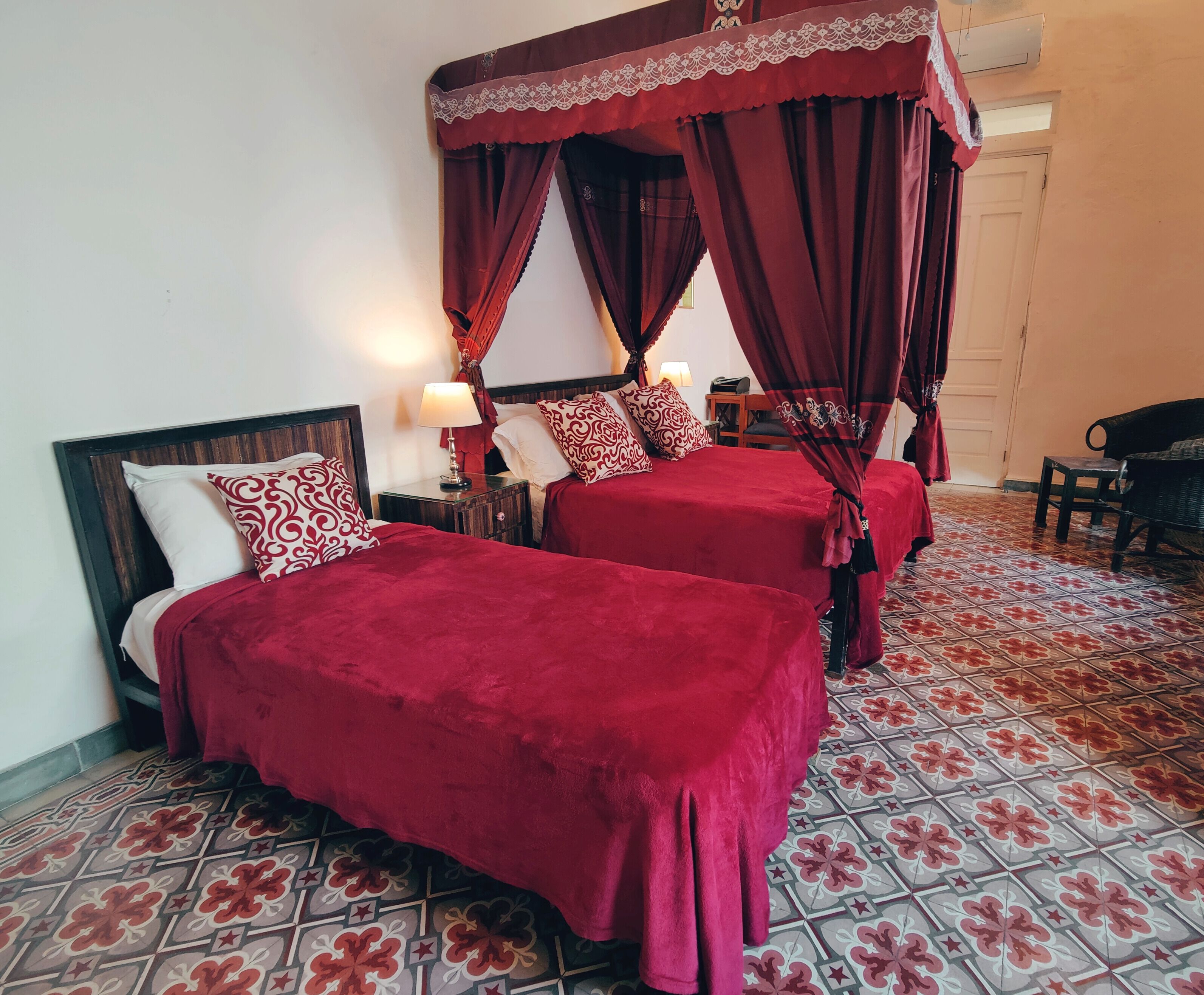 A hotel room with two beds, each with red bedding and a canopy. The room has a tiled floor with a floral pattern.