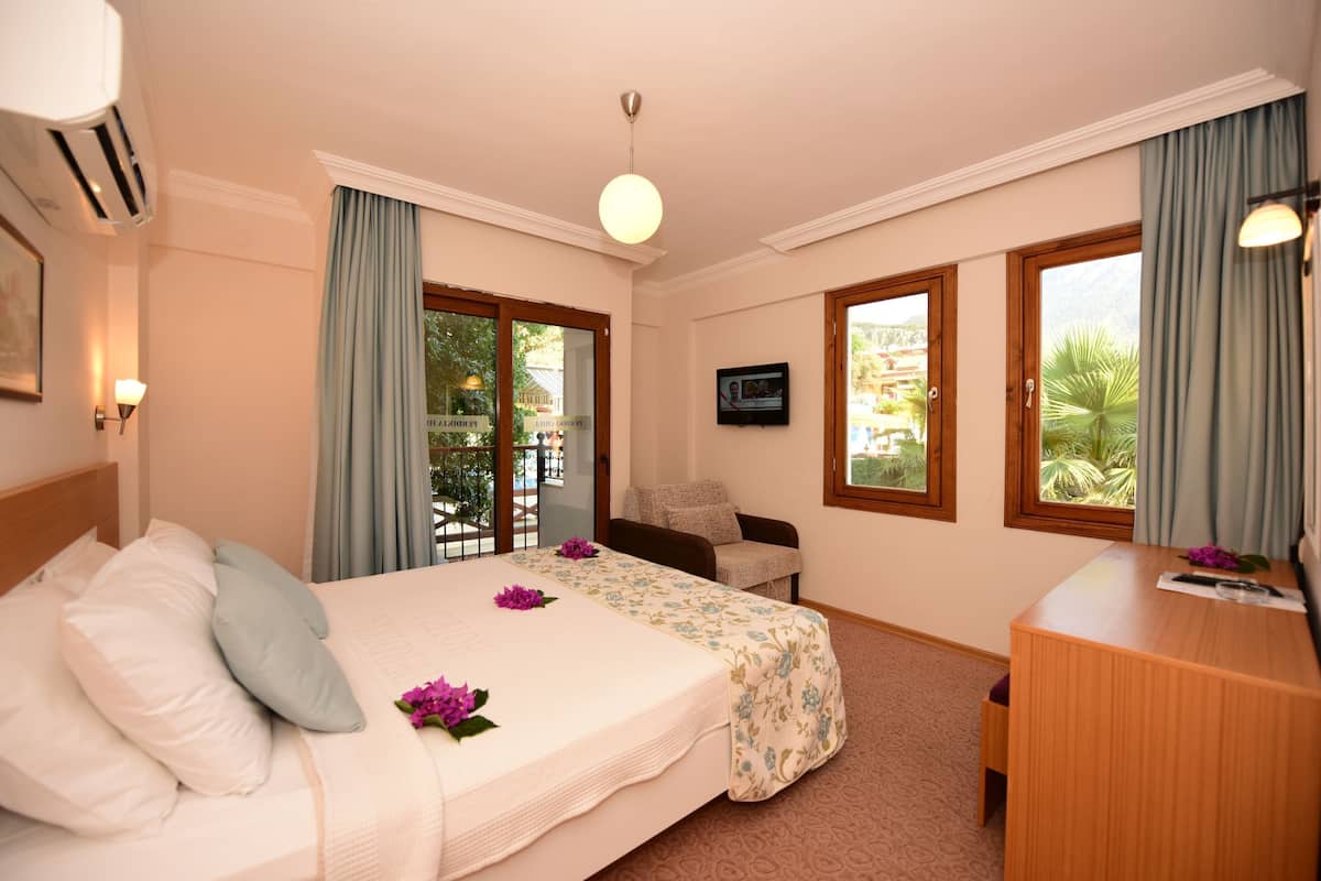 Quarto de hotel com cama, escrivaninha, cadeira, televisão e amplas janelas com cortinas.