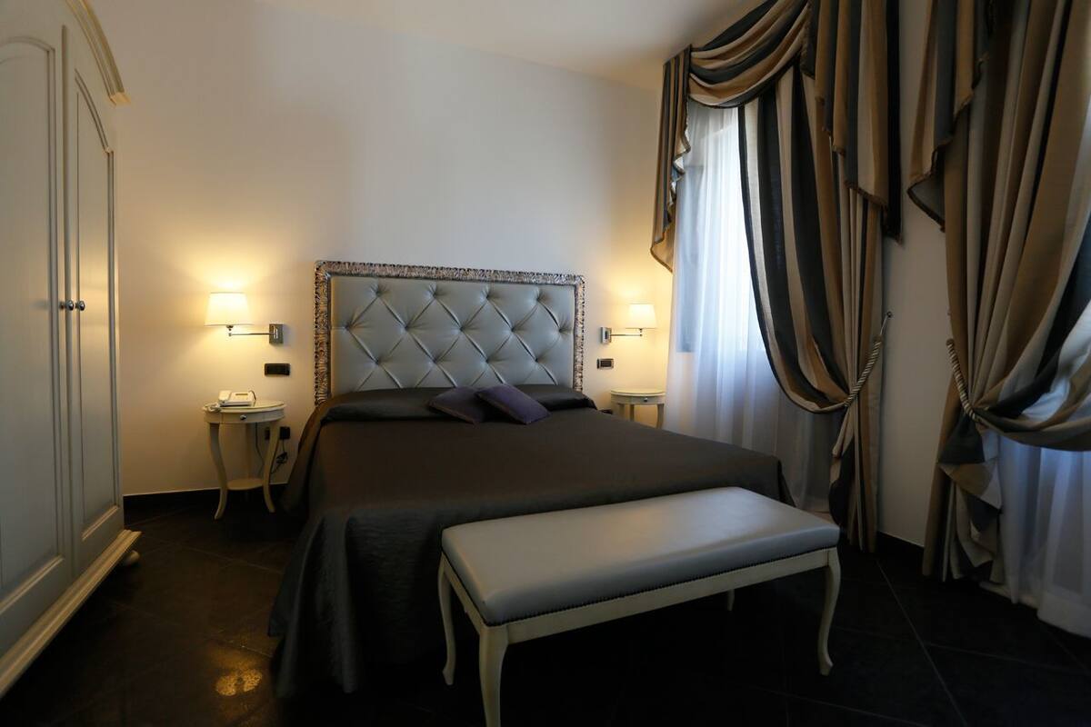 A neatly arranged bedroom with a tufted headboard, bedside tables with lamps, a bench, and a large window with curtains.
