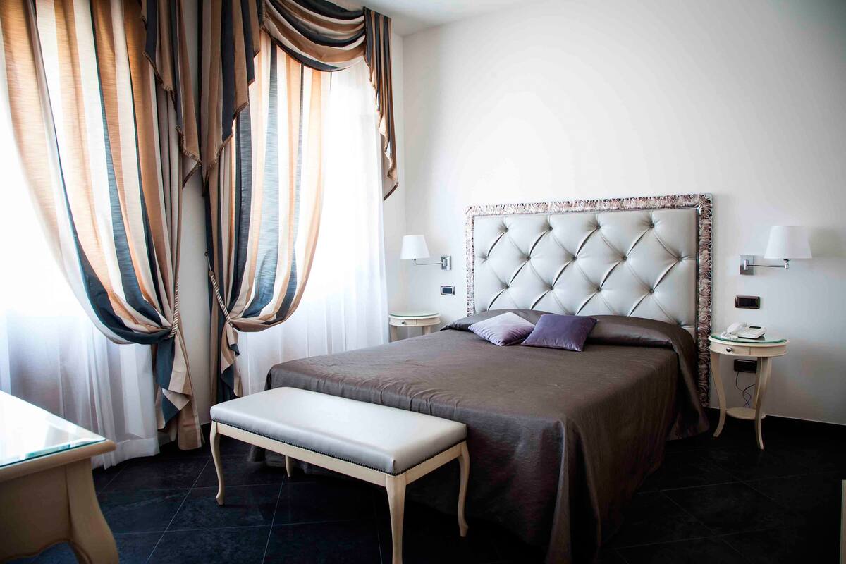 A hotel room with a large bed, a bedside table, a bench, and curtains.