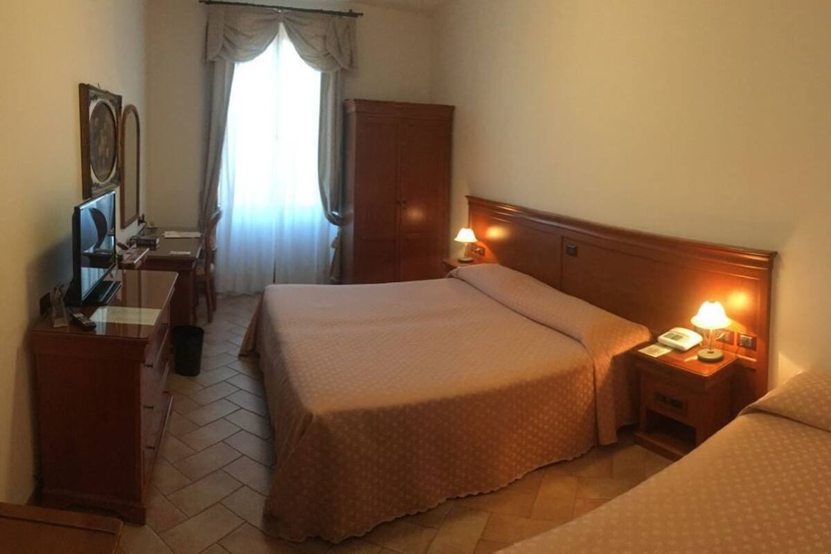 A hotel room with a large bed, a desk with a computer, a wardrobe, and a bedside table with a lamp.