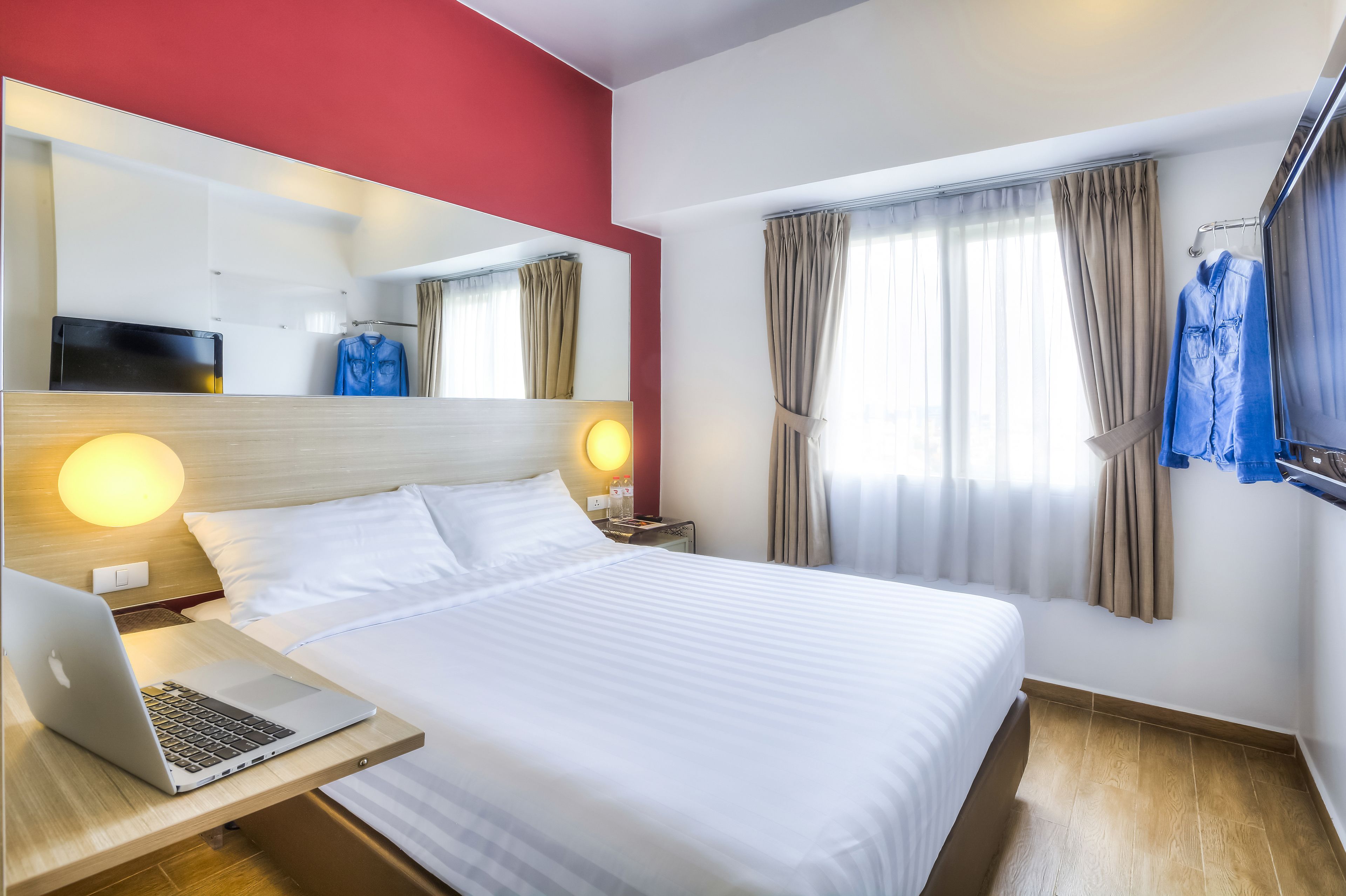 A hotel room with a bed, a desk with a laptop, a TV, and a mirror.