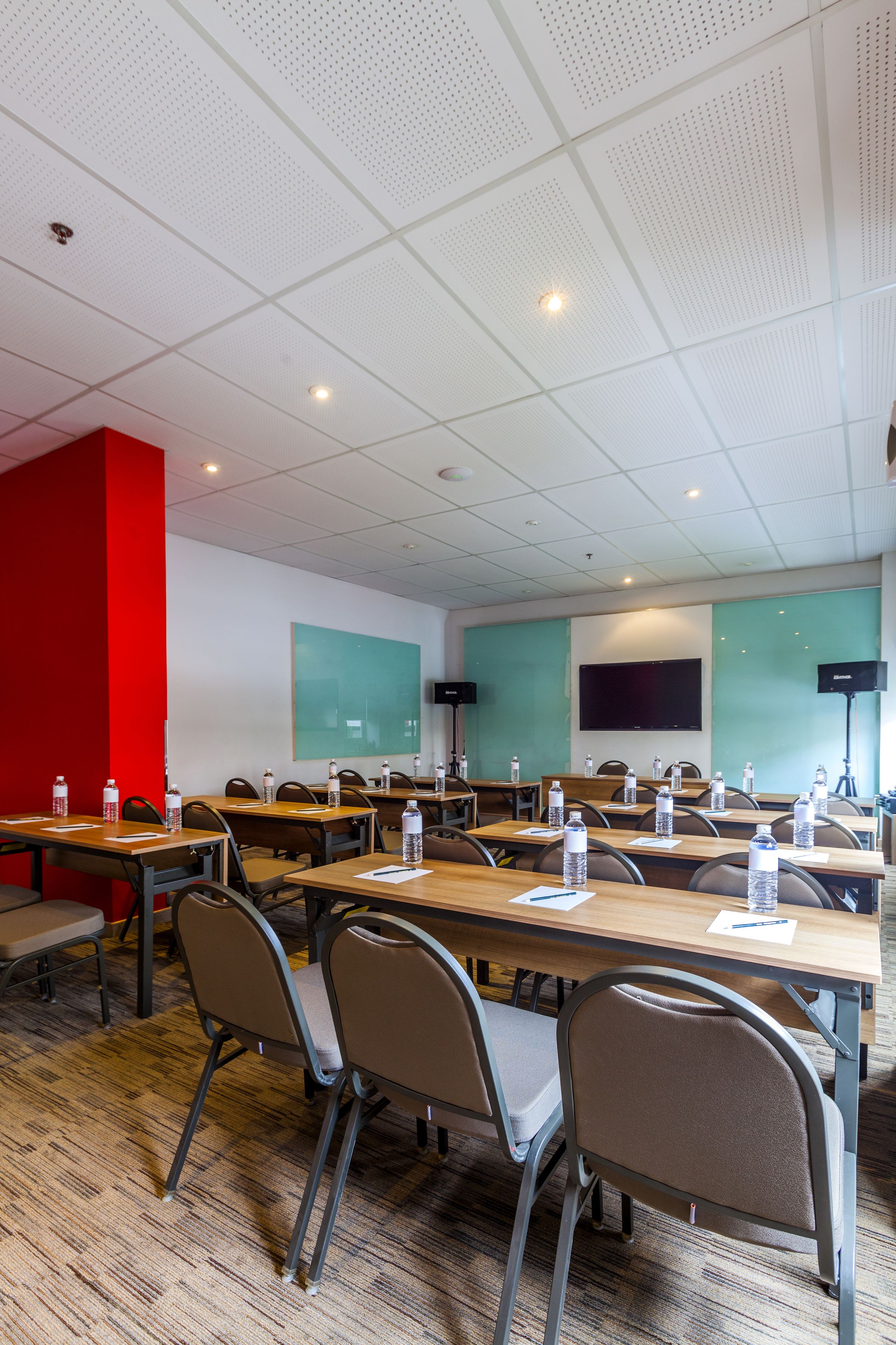 Un salón de conferencias con mesas, sillas, botellas de agua, una pared roja decorativa, una pantalla grande y un orador.