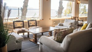 Een moderne woonkamer met een bank, fauteuils en een salontafel. Er zijn grote ramen met uitzicht op zee en palmbomen.