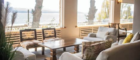 Una sala de estar moderna con un sofá, sillones y una mesa de centro. Hay ventanas amplias con vistas al mar y palmeras.