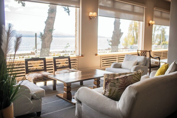 Una sala de estar moderna con un sofá, sillones y una mesa de centro. Hay ventanas amplias con vistas al mar y palmeras.