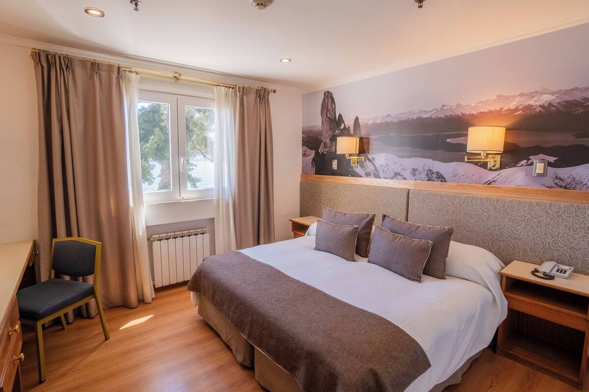 Een hotelkamer met een groot bed, een stoel, een nachtkastje met een lamp, en een berglandschap op de muur.
