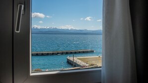Uitzicht door een raam met een metalen hek, een wateroppervlak, een steiger en besneeuwde bergen in de verte.