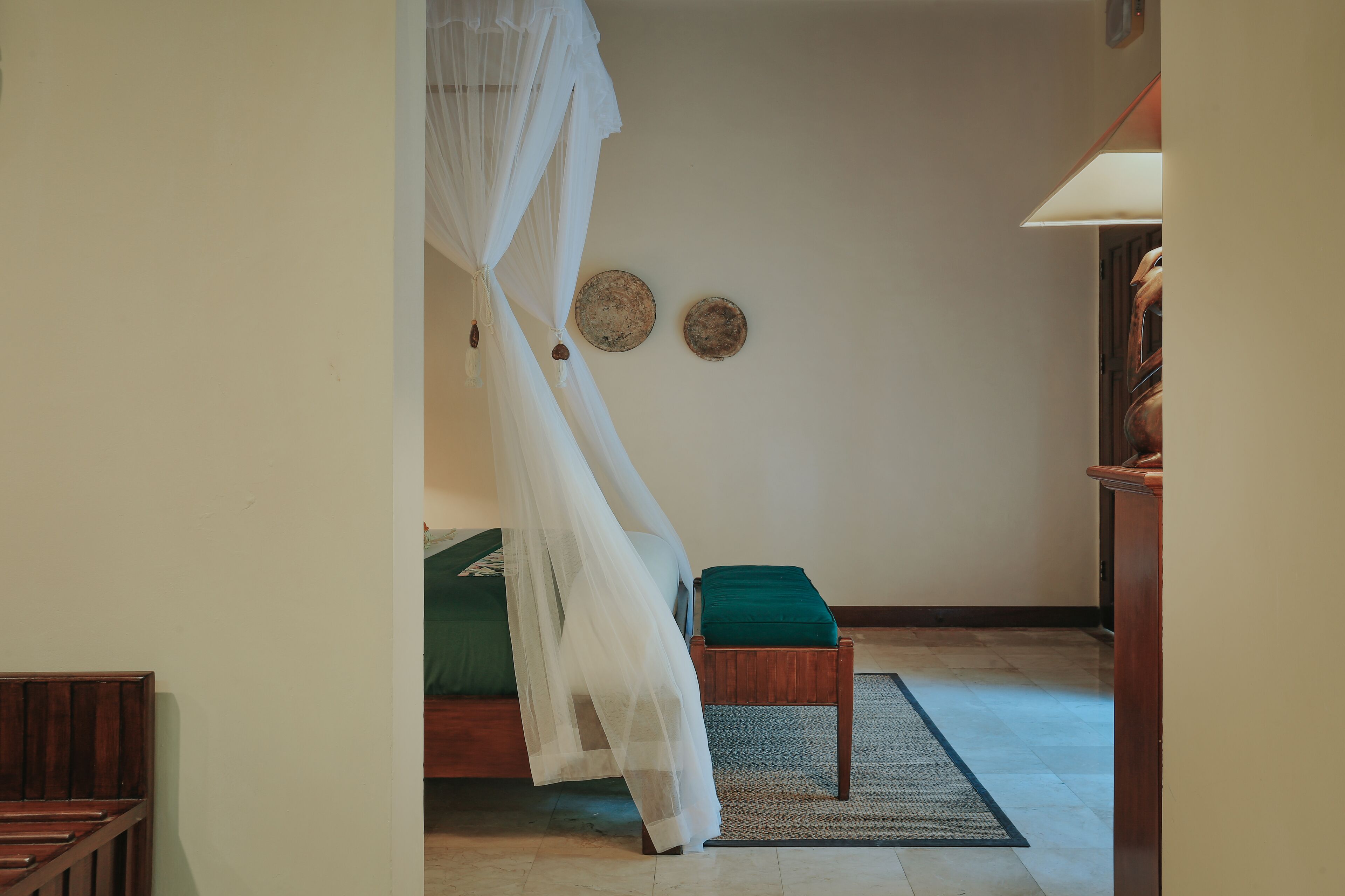 Un dormitorio con una cama, un banco de madera, un cojín verde, un dosel blanco y dos platos colgados en la pared.