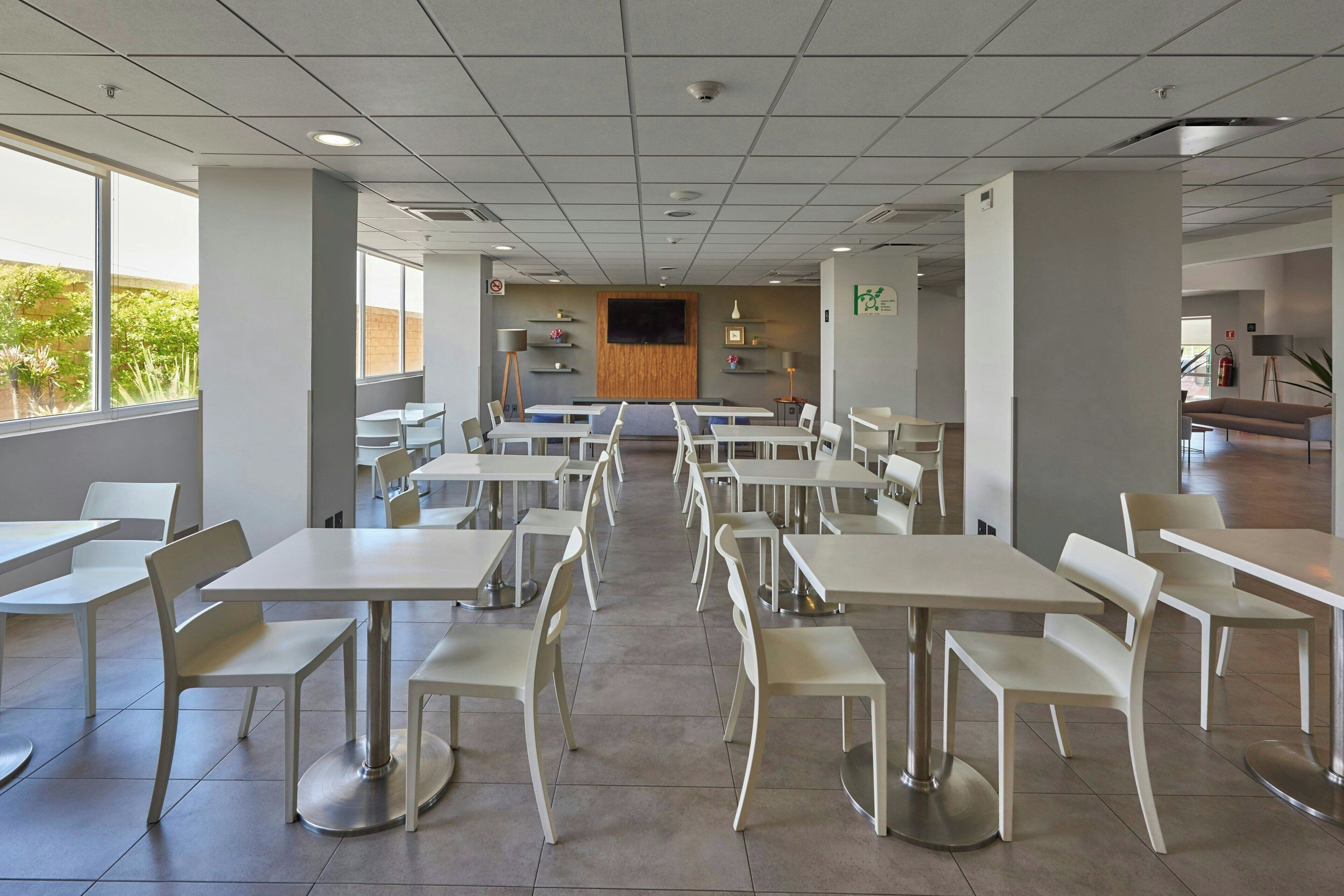 Una amplia cafetería con varias mesas y sillas, ventanas grandes y una pantalla de televisión central.