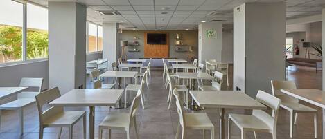 A spacious cafeteria with multiple tables and chairs, large windows, and a central television screen.