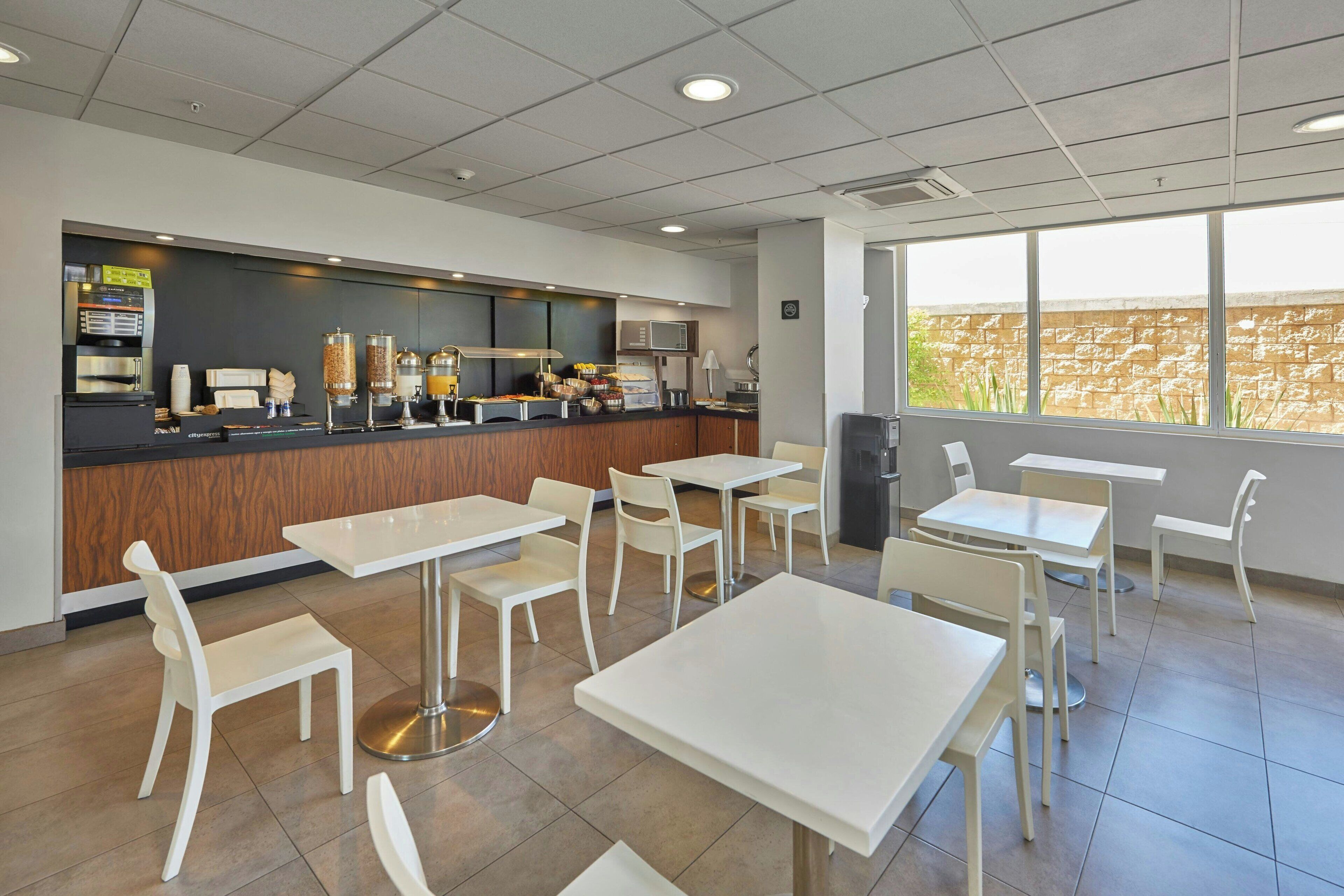 Un café moderne avec des tables et des chaises blanches, un appareil à café et une vue sur un mur de briques à travers de grandes fenêtres.